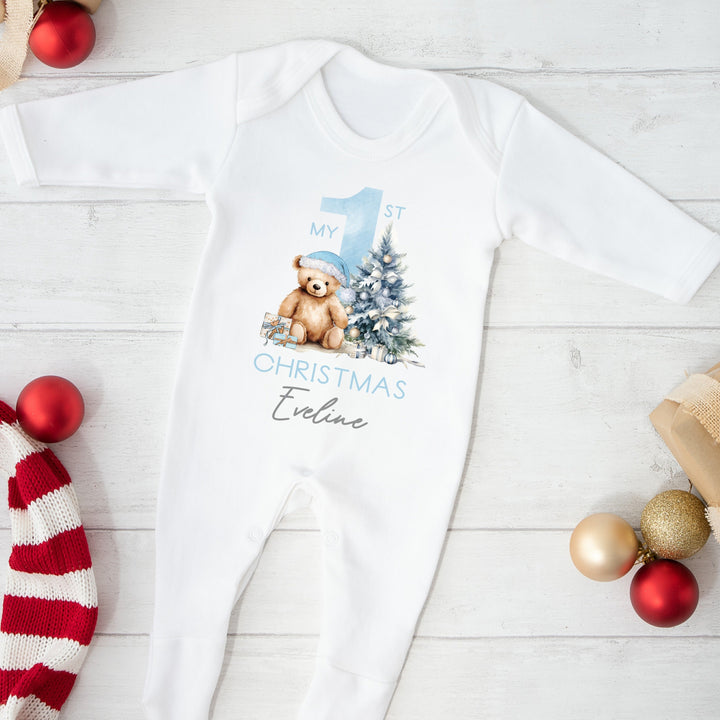 A white baby grow that features a big blue 1 and a teddy bear wearing a blue christmas hat sitting next to a christmas tree.