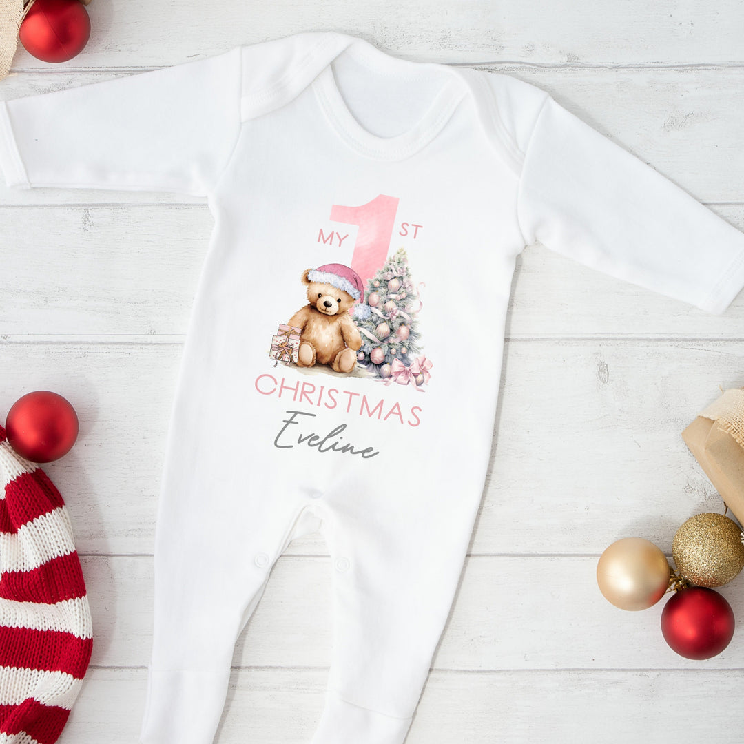 A white baby grow that features a big pink 1 and a teddy bear wearing a pink christmas hat sitting next to a christmas tree.