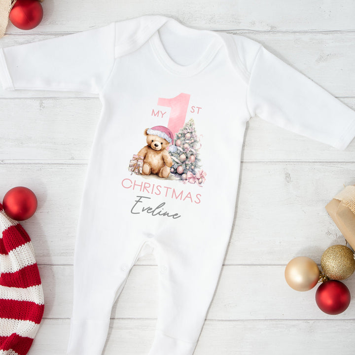 A white baby grow that features a big pink 1 and a teddy bear wearing a pink christmas hat sitting next to a christmas tree.
