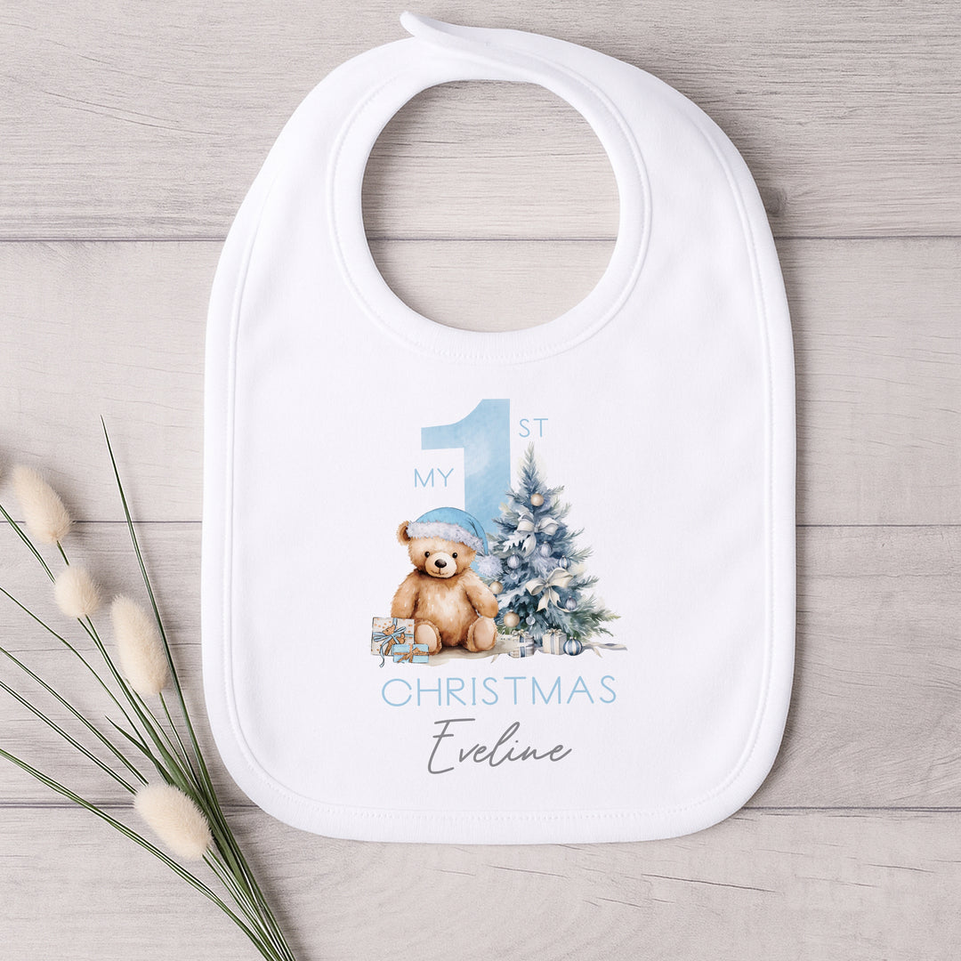 A white baby bib that features a big blue 1 and a teddy bear wearing a blue christmas hat sitting next to a christmas tree.