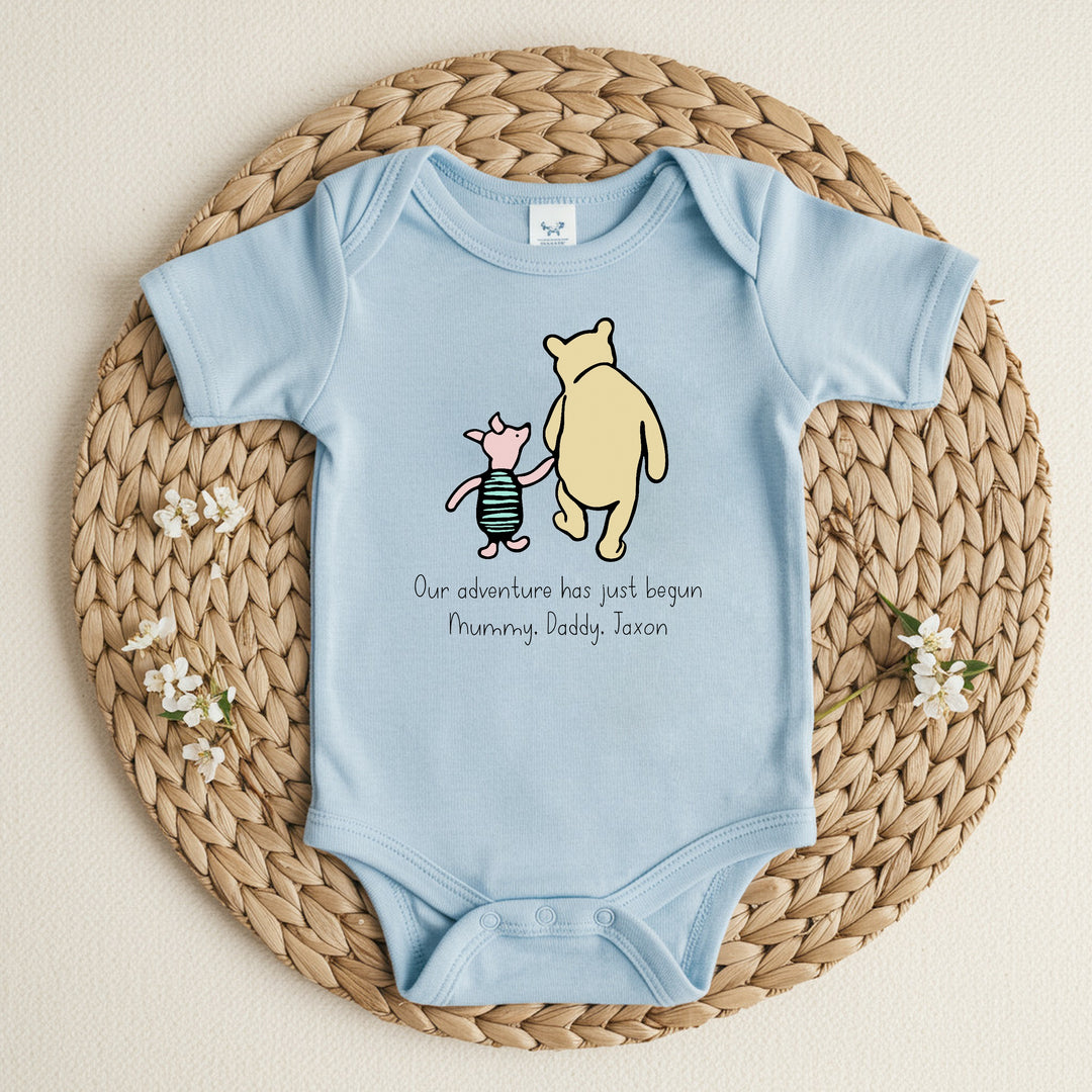 a personalised blue baby vest that says 'Our adventure has jusy begun Mummy, Daddy, Jaxon'. This design features a classic winnie the pooh and piglet