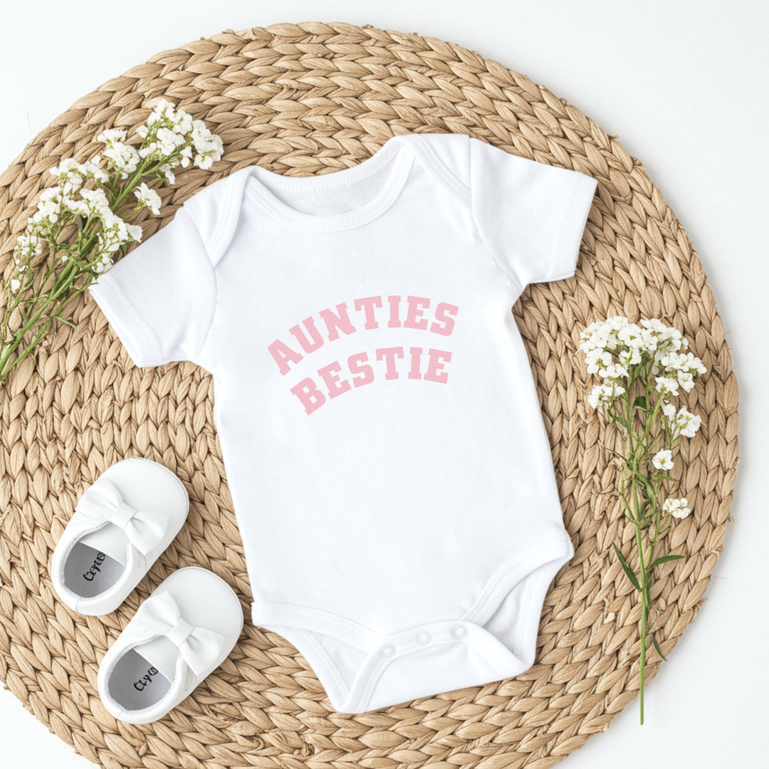 White baby onesie with 'Aunties Bestie' text, white baby shoes, and baby's breath flowers on a woven mat.