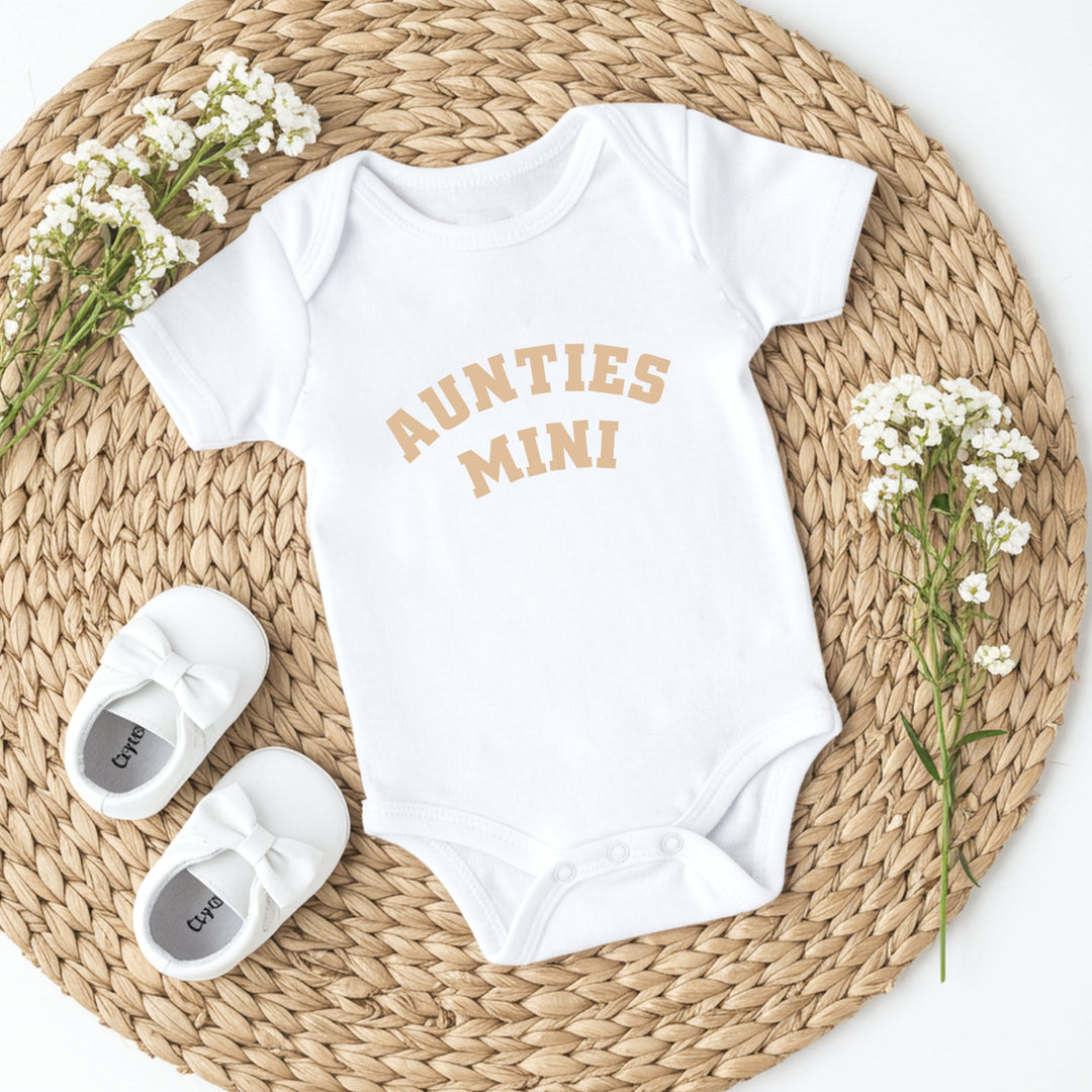 White baby onesie with 'Aunties Mini' text, white shoes, and flowers on a woven mat.