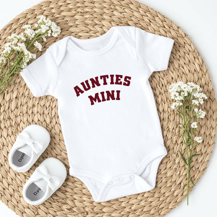 White baby onesie with 'AUNTIES MINI' text, white baby shoes, and small flowers on a woven mat.