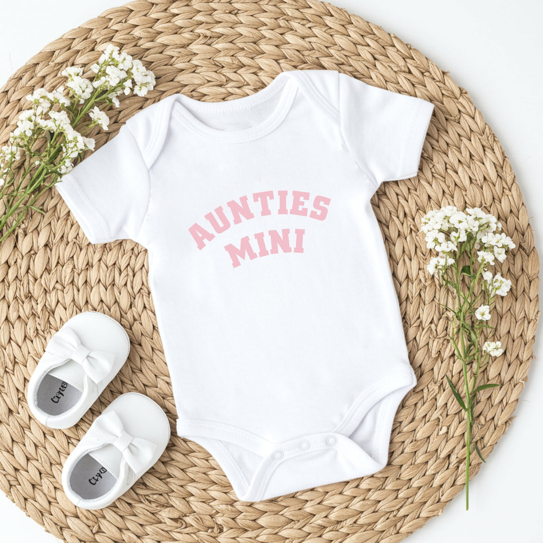 White baby onesie with 'AUNTIES MINI' text, white baby shoes, and baby flowers on a woven mat.