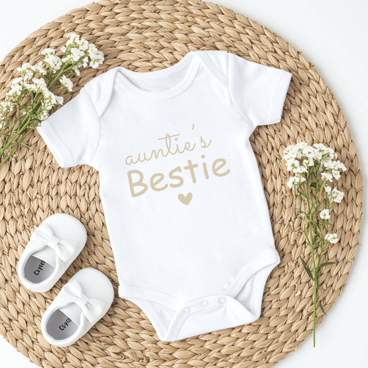 White baby onesie with 'auntie's Bestie' text, white baby shoes, and white flowers on a woven mat.
