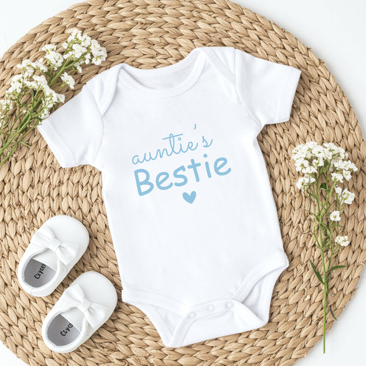White baby onesie with 'Auntie's Bestie' text, white shoes, and baby's breath flowers on a woven mat.