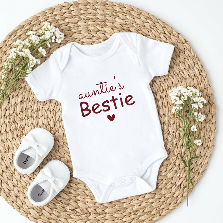 White baby onesie with 'Auntie's Bestie' text on a woven mat with baby shoes and flowers.
