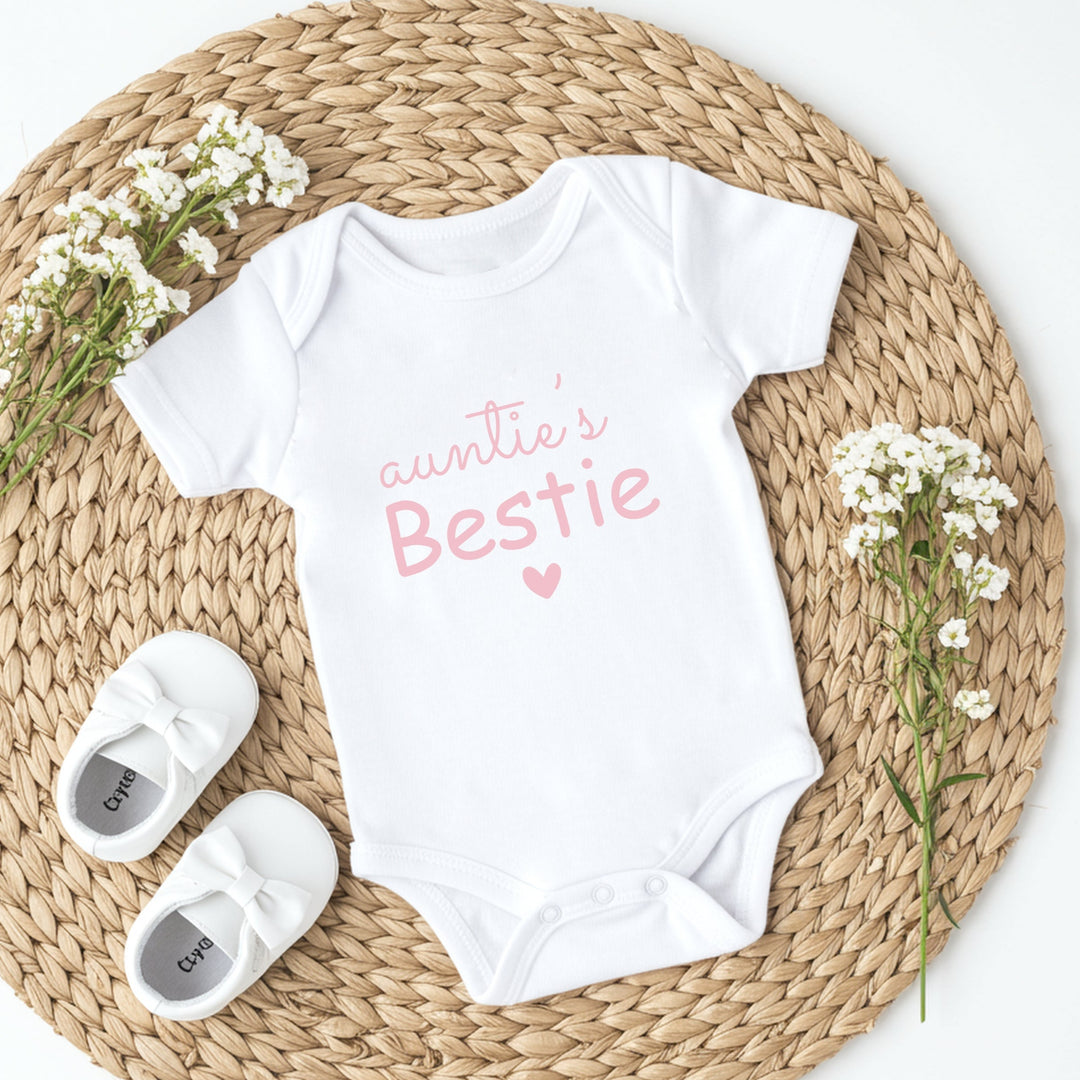 White baby onesie with 'Auntie's Bestie' text, white baby shoes, and baby flowers on a woven mat.