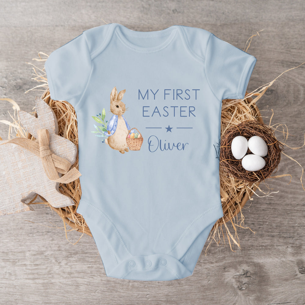 Personalised blue Easter baby vest that says 'My First Easter Oliver'. This design features a rabbit wearing a blue jacket holding an Easter basket