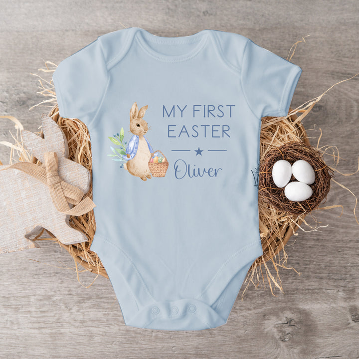 Personalised blue Easter baby vest that says 'My First Easter Oliver'. This design features a rabbit wearing a blue jacket holding an Easter basket
