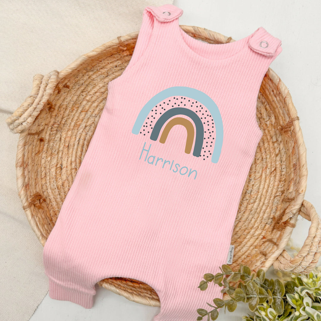 Personalised pink ribbed dungarees that say 'Harrison'. This design features a blue rainbow