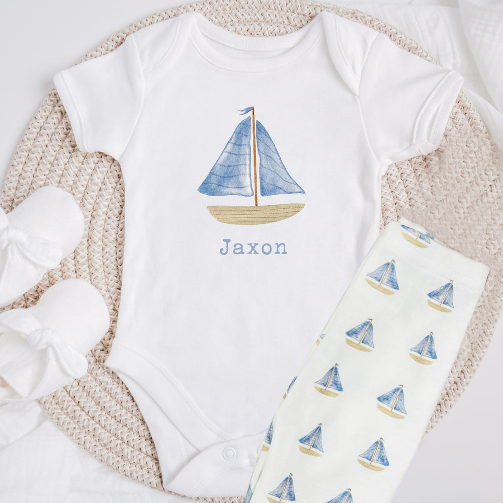 a personalised white baby vest that says 'Jaxon'. This design features a blue sail boat