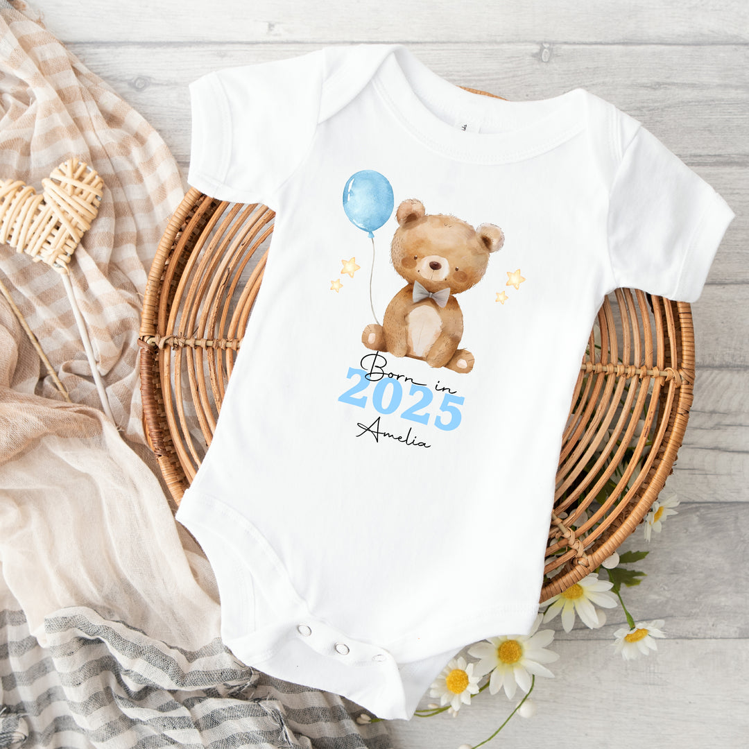 a personalised white baby vest that says 'Born in 2025 Amelia'. This design features a brown teddy holding a blue balloon