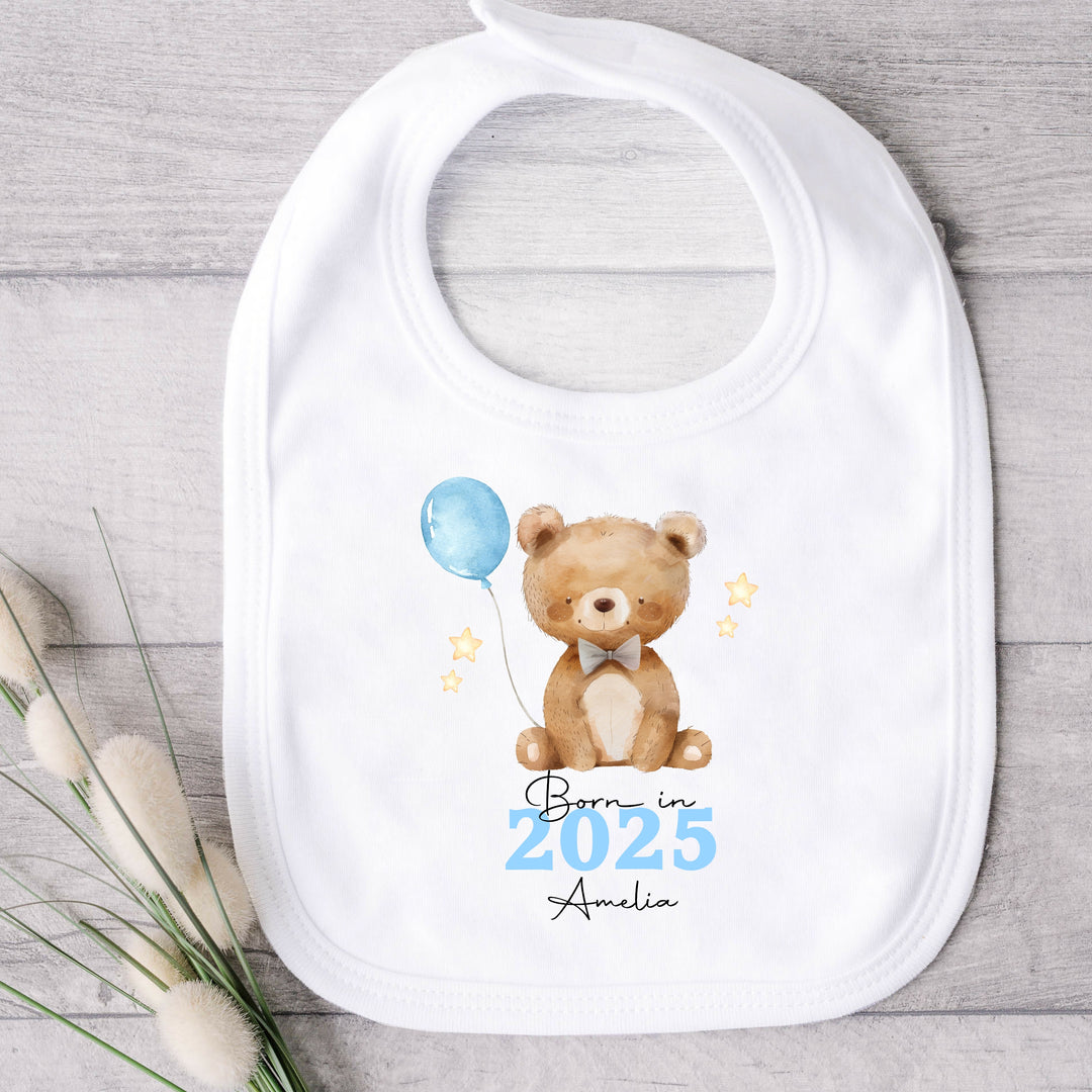 a personalised white baby bib that says 'Born in 2025 Amelia'. This design features a brown teddy holding a blue balloon