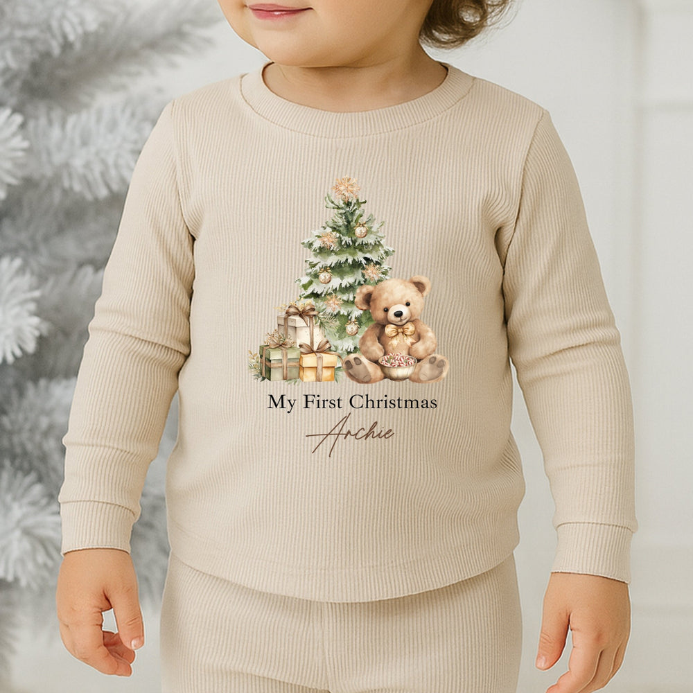 Child wearing a beige outfit with a Christmas design in front of a decorated tree.