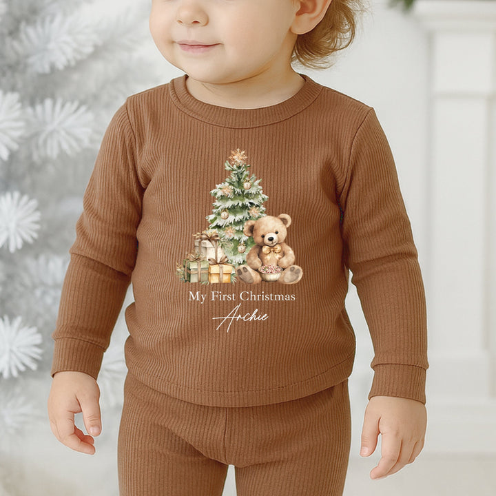Child wearing a brown Christmas-themed outfit with a tree and teddy bear design, standing against a snowy background.