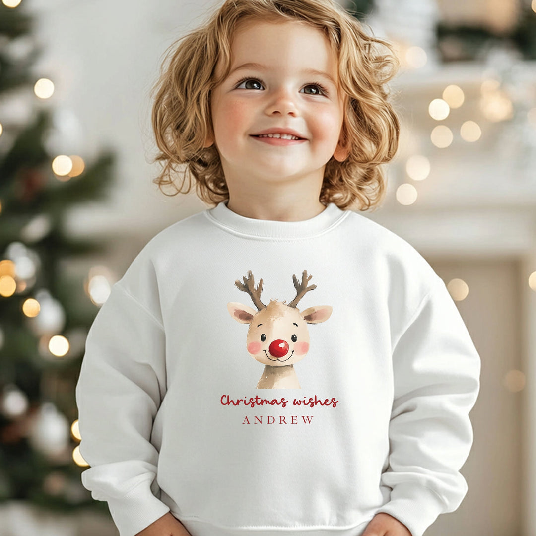 Child wearing a white sweatshirt with a reindeer design and 'Christmas wishes ANDREW' text, standing in a festive setting.