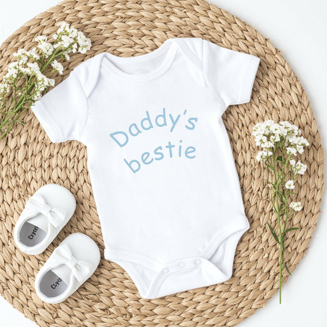 White baby onesie with 'Daddy's bestie' text, white baby shoes, and baby's breath flowers on a woven mat.