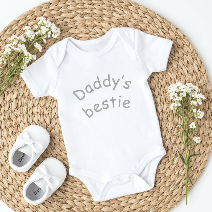 White baby onesie with 'Daddy's bestie' text, white baby shoes, and baby's breath flowers on a woven mat.