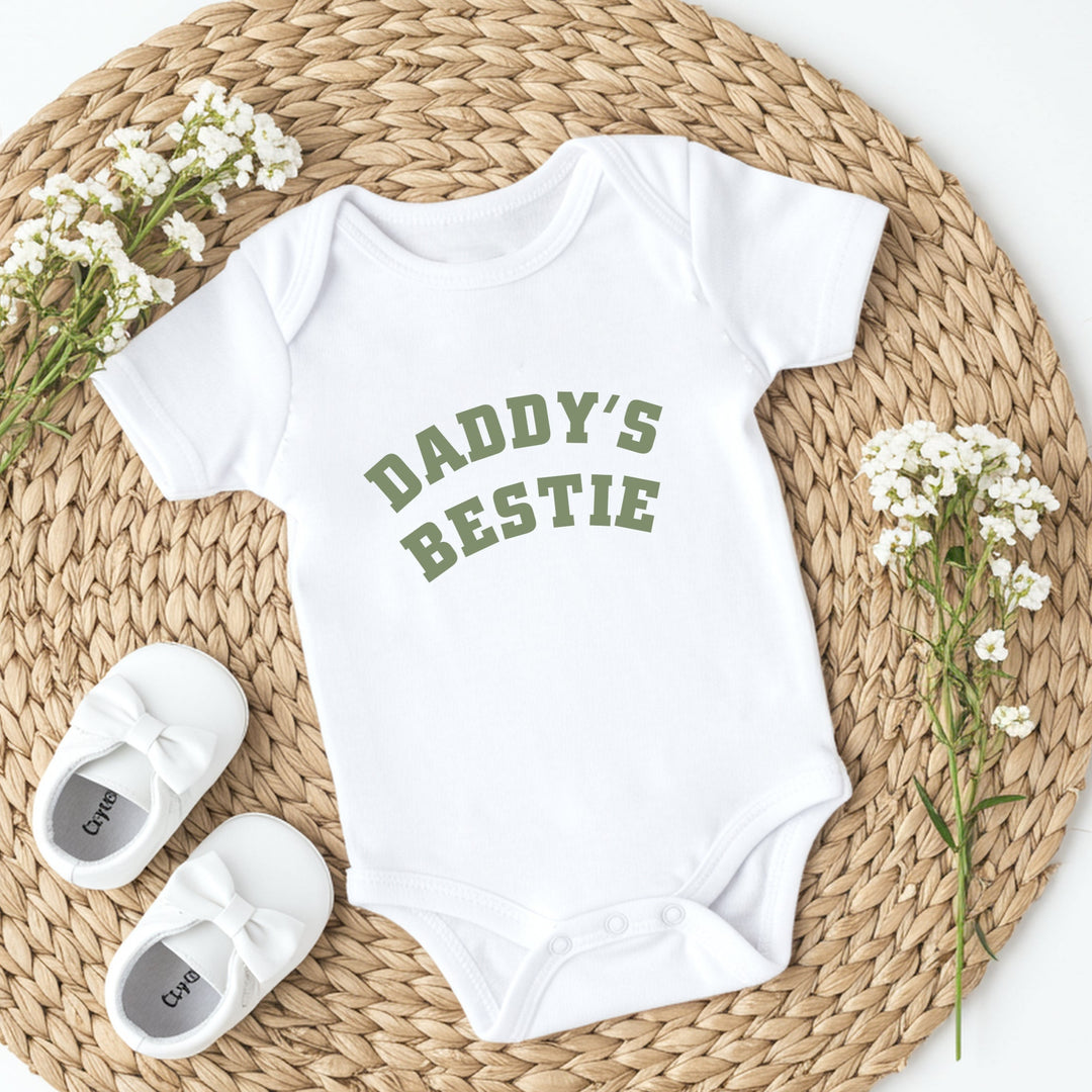 White baby onesie with 'Daddy's Bestie' text, white baby shoes, and baby's breath flowers on a woven mat.