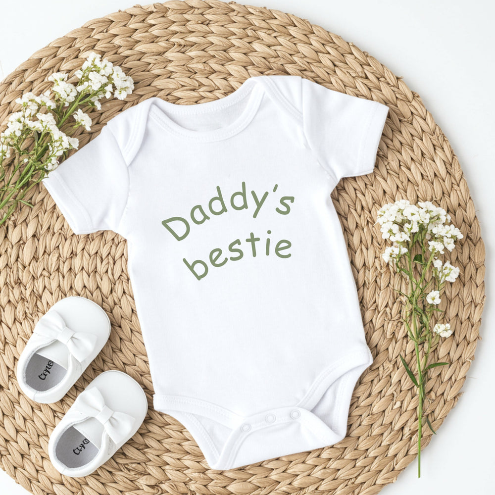 White baby onesie with 'Daddy's bestie' text, white baby shoes, and baby's breath flowers on a woven mat.