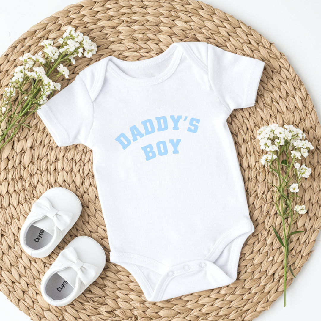 White baby onesie with 'Daddy's Boy' text, white baby shoes, and baby's breath flowers on a woven mat.