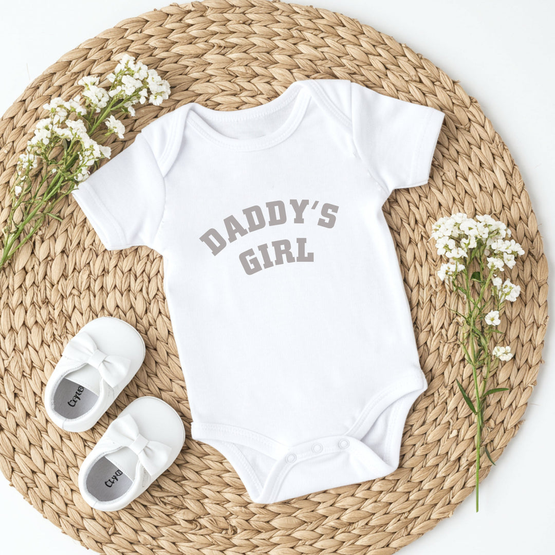 White baby onesie with 'Daddy's Girl' text, white baby shoes, and baby's breath flowers on a woven mat.