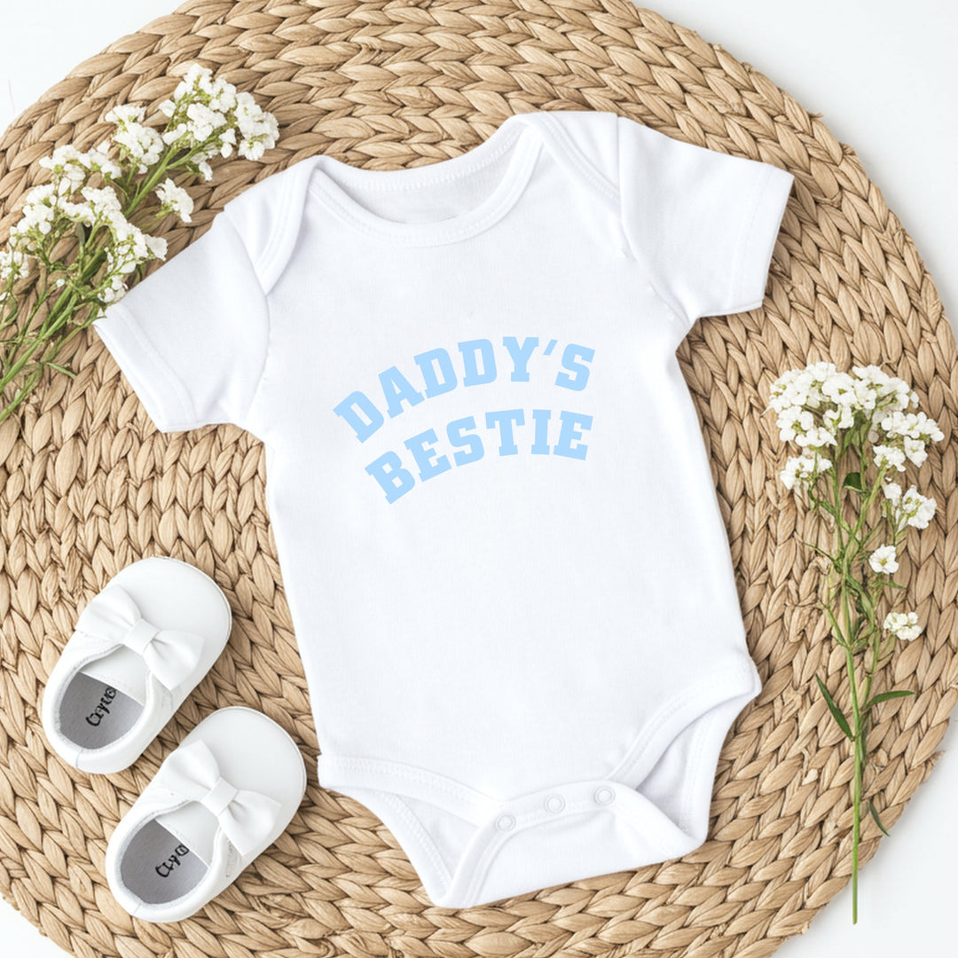 White baby onesie with 'Daddy's Bestie' text, white baby shoes, and baby's breath flowers on a woven mat.