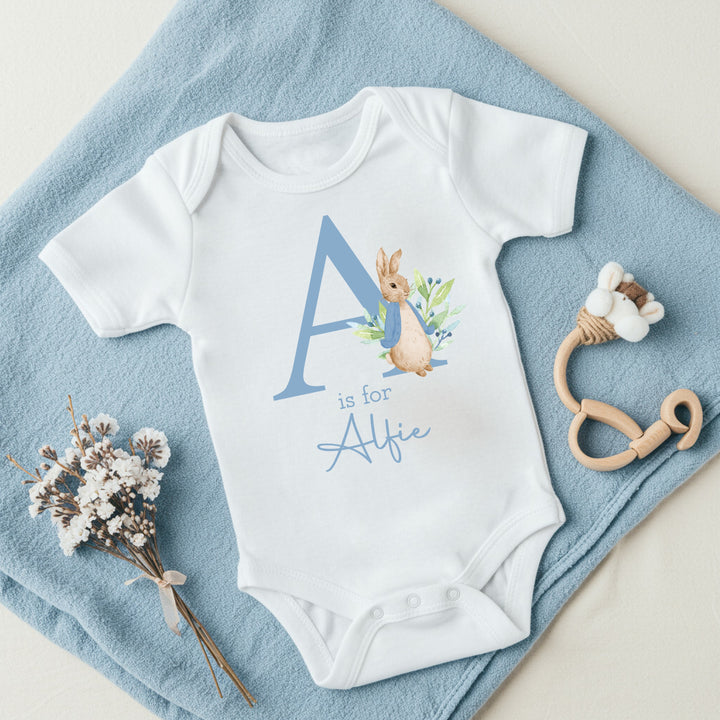 a personalised white baby vest that says 'A is for Alfie' in dusky blue text. This design features a rabbit wearing a dusky blue jacket
