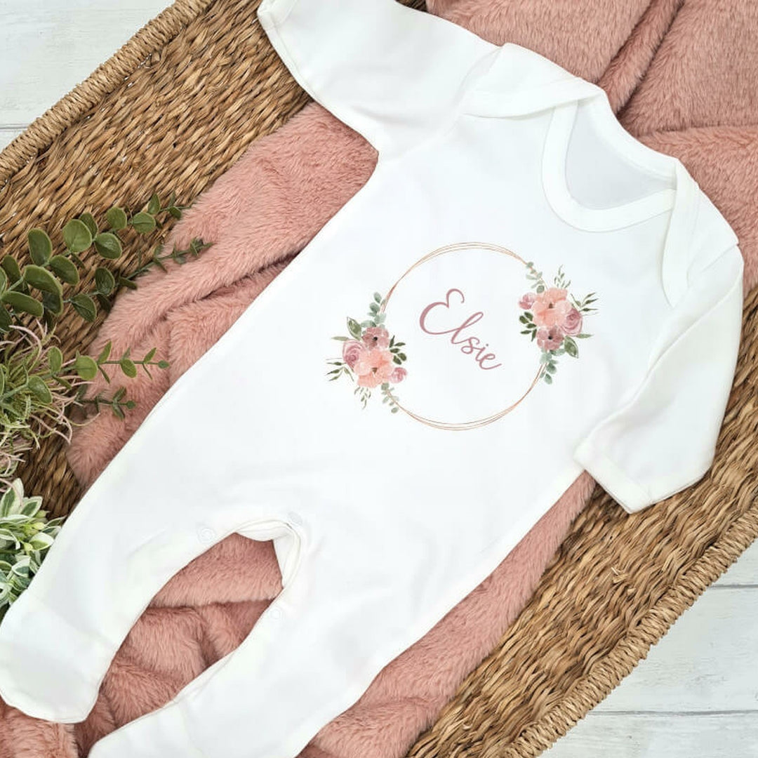 A white babygrow printed with a dusky pink gold ring with floral accents. A name can be personalised in the center of the ring.