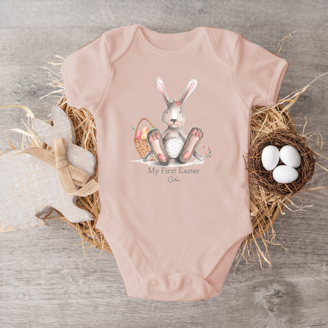 Personalised Pink Easter baby vest that says 'My First Easter Cole'. This design features a grey rabbit with pink details sitting next to an Easter basket with eggs in it