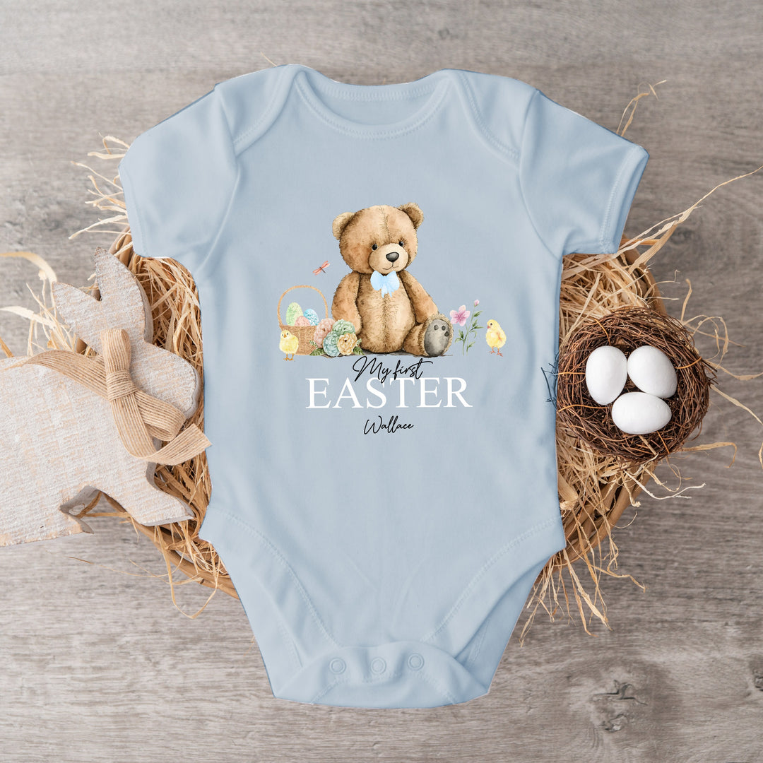 Personalised blue Easter baby vest that says 'My First Easter Wallace'. This design features a teddy bear wearing a blue bow tie sitting next to an Easter basket and 2 chicks