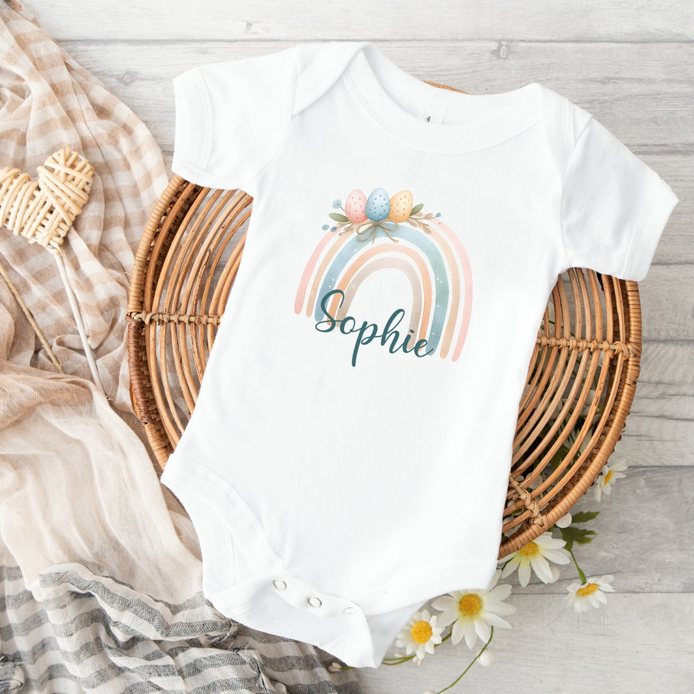 Personalised white Easter baby vest that says 'Charlie'. This design features a pastel rainbow with Easter eggs