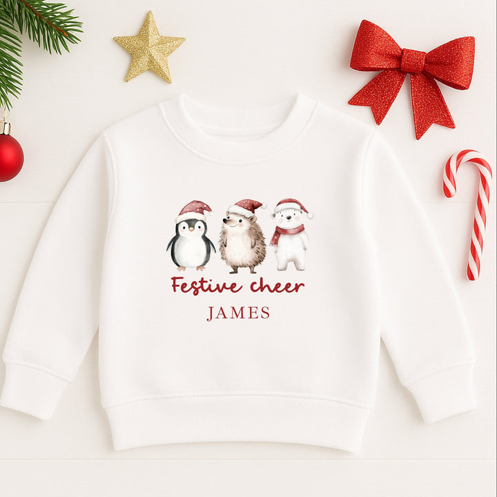 White hoodie with Christmas-themed design featuring penguin, hedgehog, and dog with text 'Festive cheer JAMES' on a white background with festive decorations.