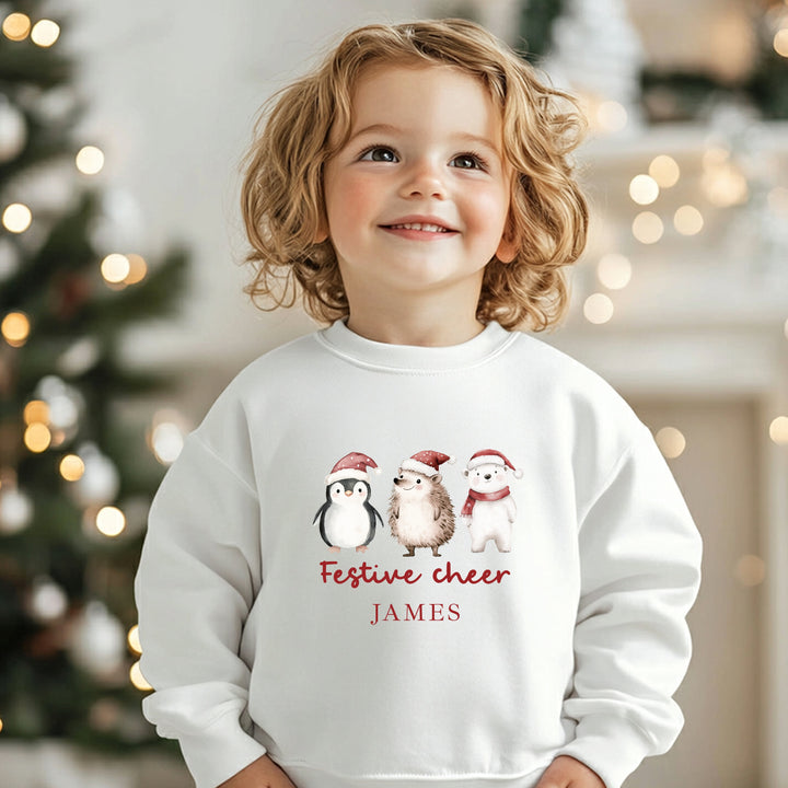 Child wearing a white sweatshirt with Christmas-themed design and text, standing in front of a decorated Christmas tree.