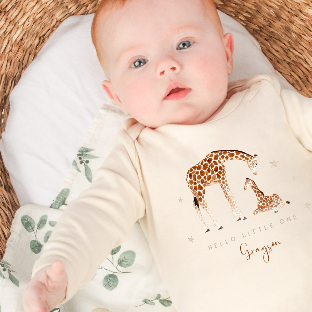 A beige babygrow featuring a design with a parent and child giraffe. The text reads "Hello little one" and has a personalised name underneath.