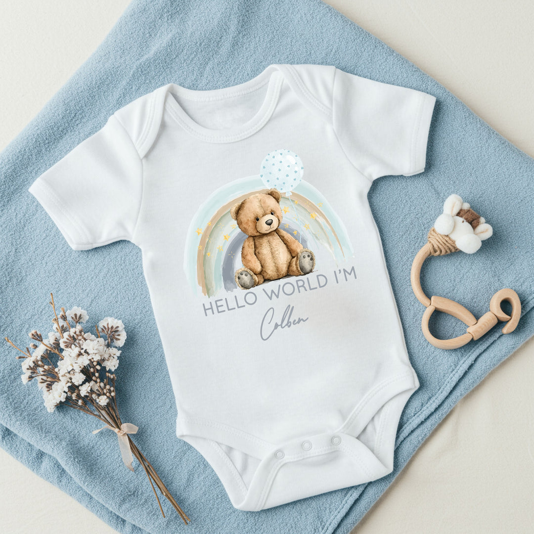 a personalised white baby vest that says 'Hello world I'm Colben'. This design features a blue rainbow with a teddy sitting in front of it holding a blue balloon