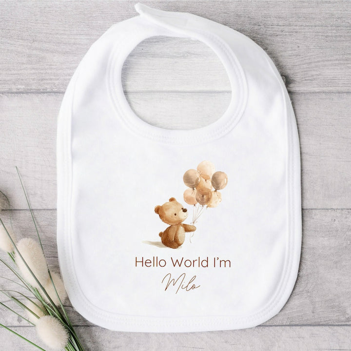 A personalised white baby bib that says 'Hello World I'm Milo'. This design features a little brown bear holding a bunch of balloons