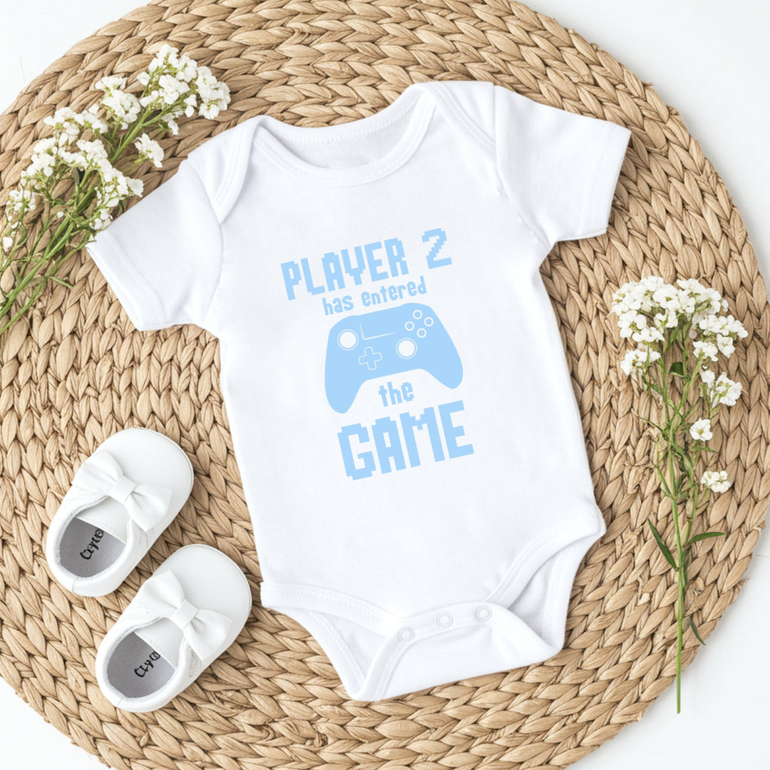 White baby onesie with 'Player 2 has entered the game' text and blue graphics on a woven mat with white shoes and flowers.