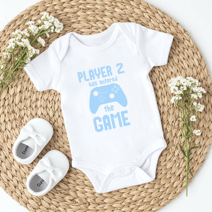 White baby onesie with 'Player 2 has entered the game' text and blue graphics on a woven mat with white shoes and flowers.