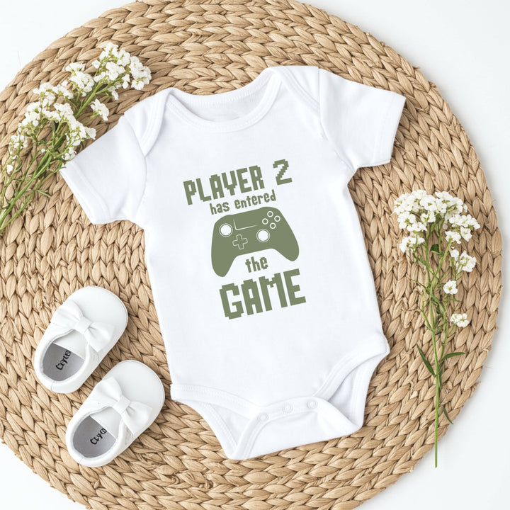 Baby onesie with 'Player 2 has entered the game' text on a woven mat with baby shoes and flowers.
