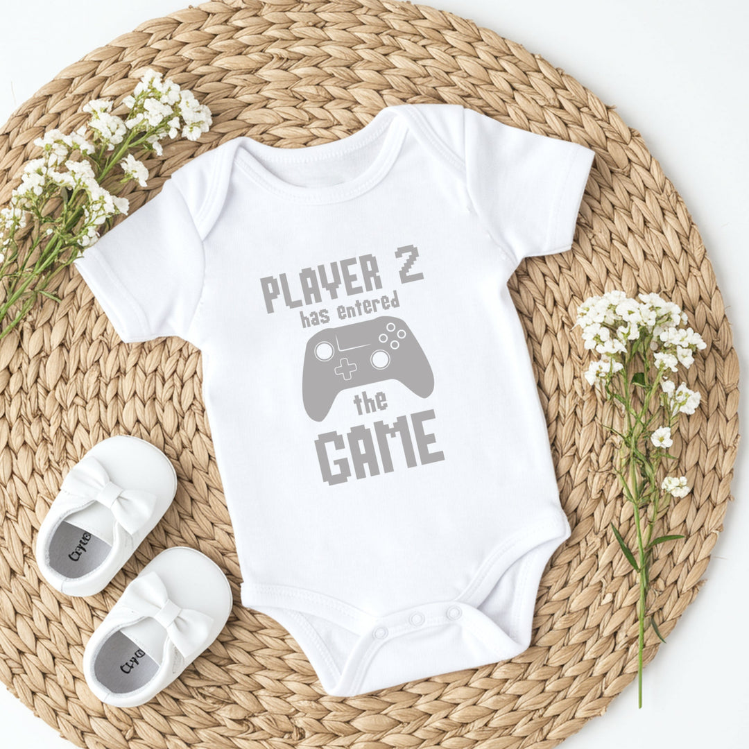 White baby onesie with 'Player 2 has entered the game' text on a woven mat with baby shoes and flowers.