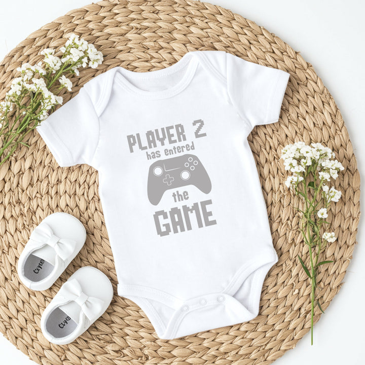 White baby onesie with 'Player 2 has entered the game' text on a woven mat with baby shoes and flowers.