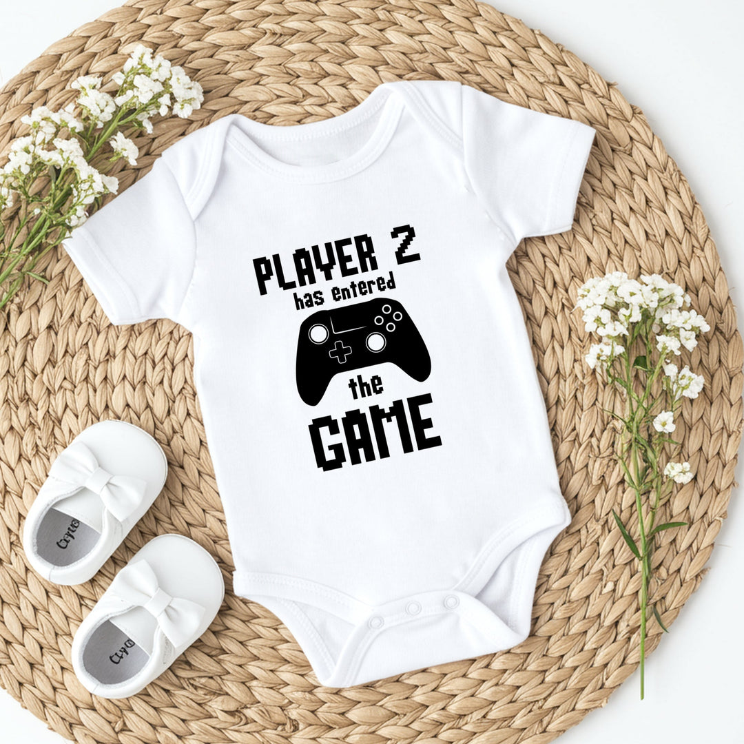 White baby onesie with 'Player 2 has entered the game' text and a black controller graphic on a woven mat with white flowers.