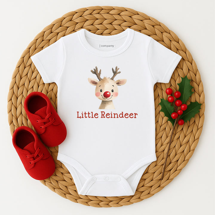 White baby onesie with reindeer design and 'Little Reindeer' text on a woven mat with red shoes and holly.