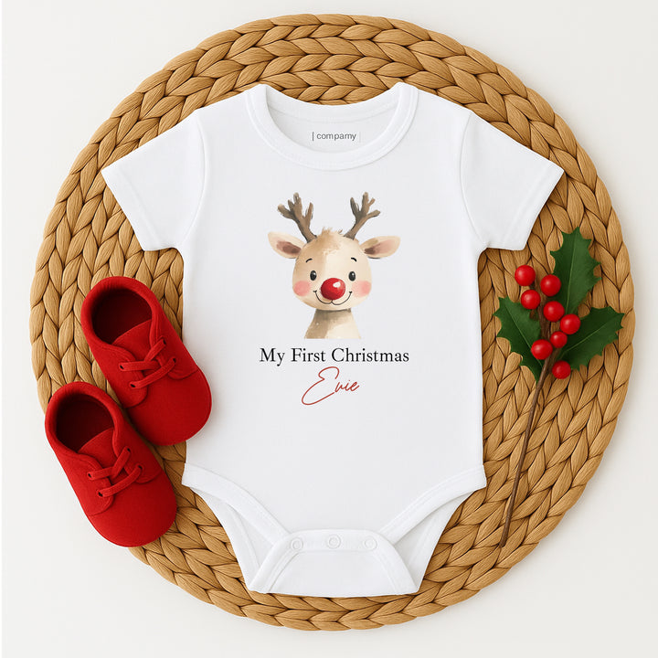 White baby onesie with reindeer design and red shoes on a woven mat
