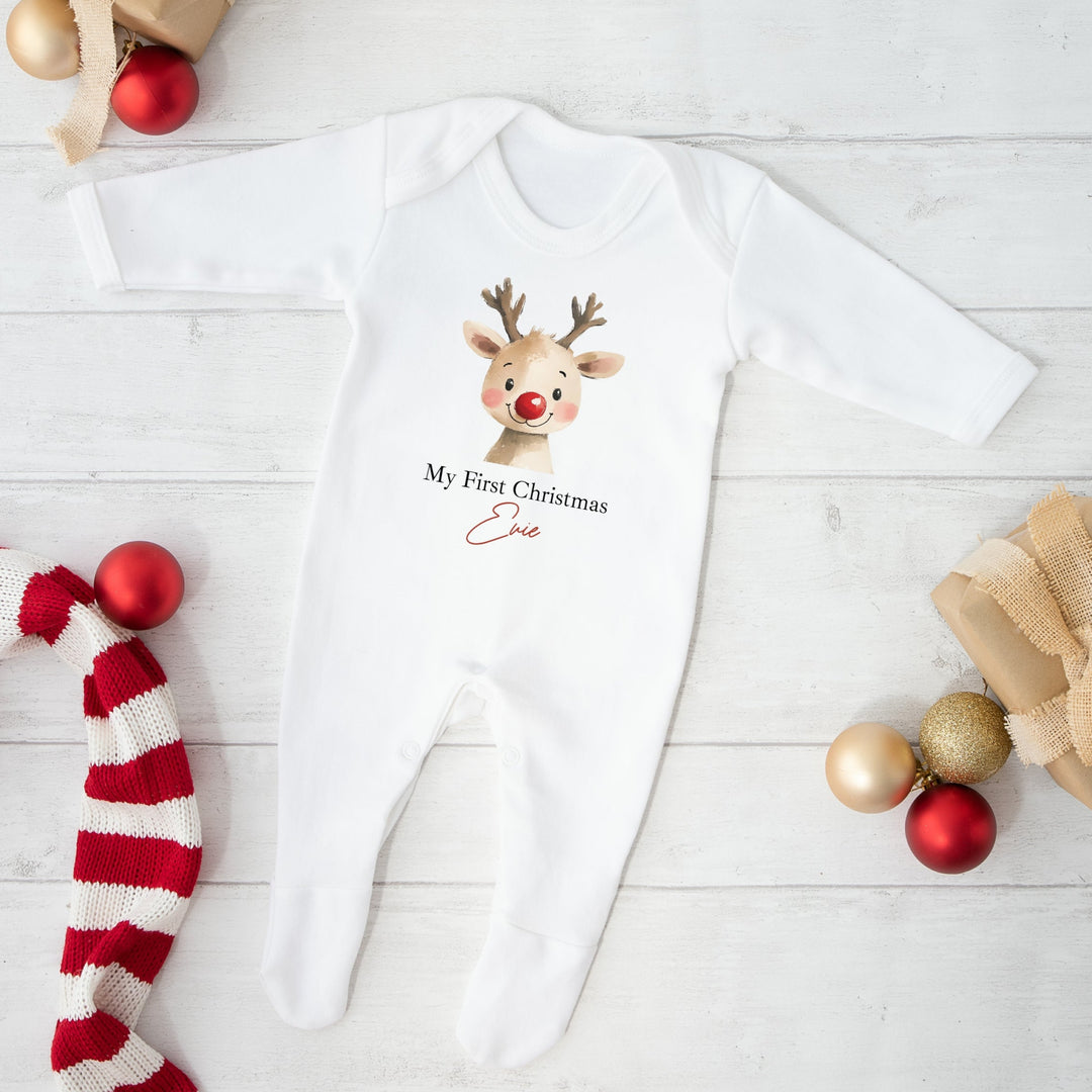 White baby onesie with reindeer design and text, surrounded by Christmas decorations on a light wooden floor.