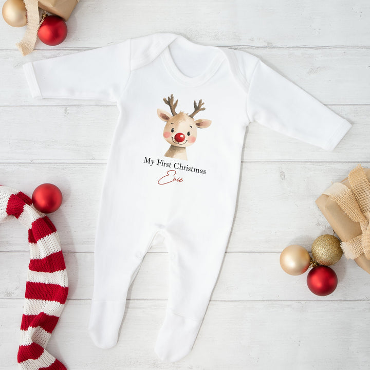 White baby onesie with reindeer design and text, surrounded by Christmas decorations on a light wooden floor.