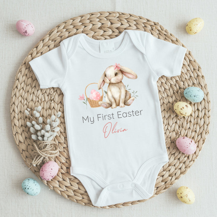 Personalised white Easter baby vest that says 'My First Easter Olivia'. This design features a brown bunny wearing a pink bow sitting next to a basket with pink eggs in it